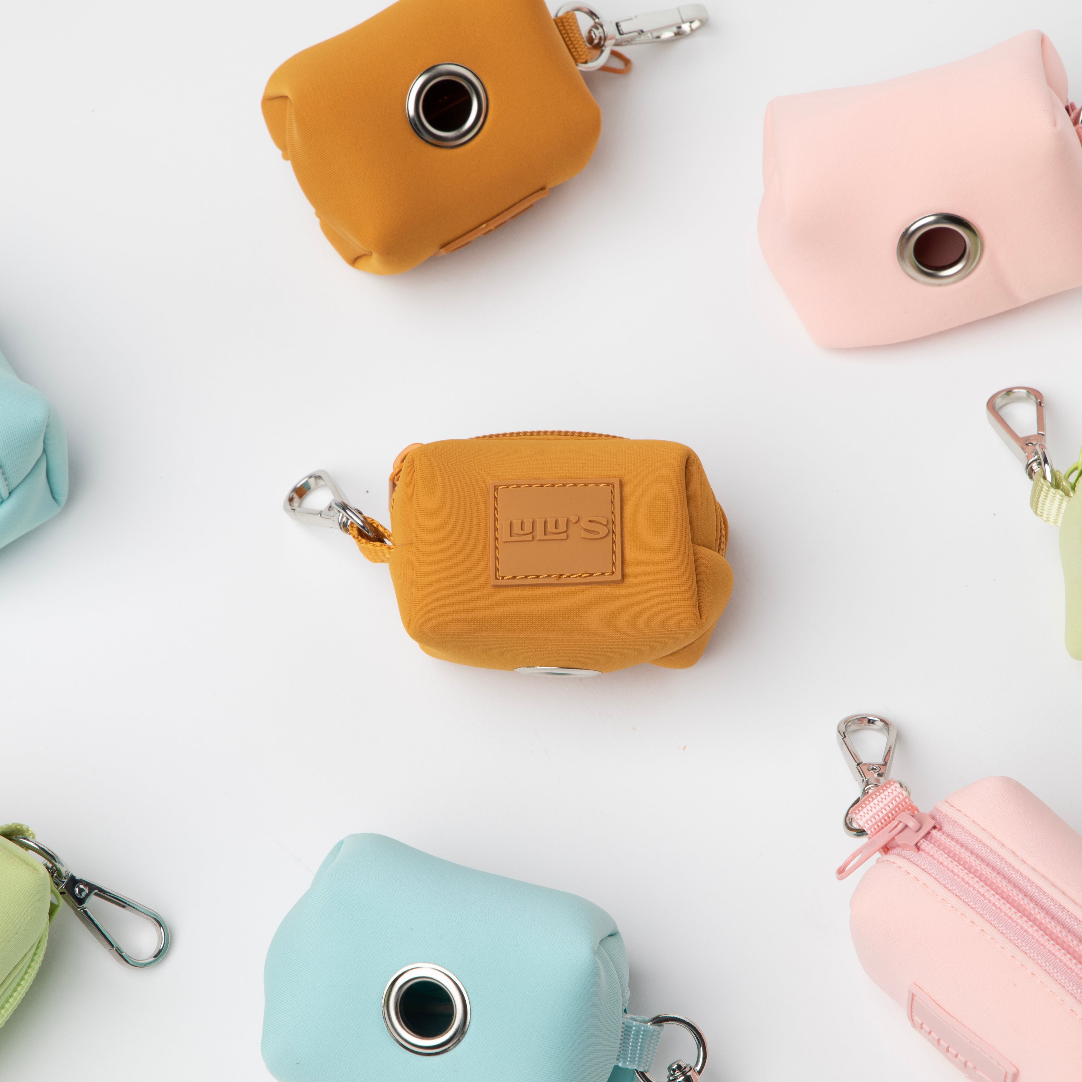 Assorted dog waste bag holders in various colors including Sunburnt Orange, Nosey Pink, Citrus Green, and Cali Blue, displayed on a white background.