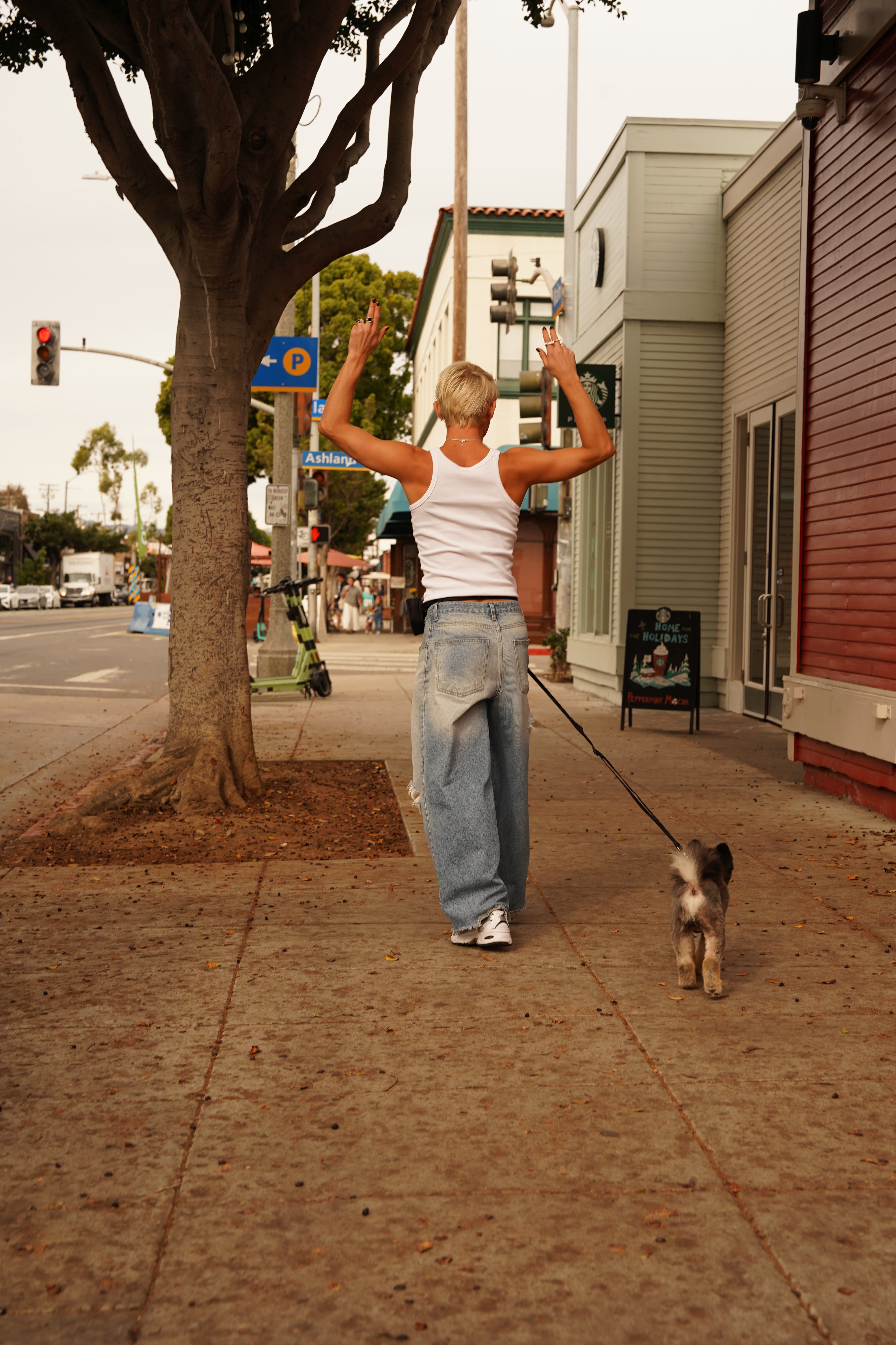 Person dancing joyfully on a city sidewalk while walking a small dog, wearing relaxed blue jeans and a white tank top, using Lulu's from Cali hands-free leash and pouch set in the color midnight black. The scene captures a lively moment with tree-lined streets, storefronts, and traffic signs in the background.