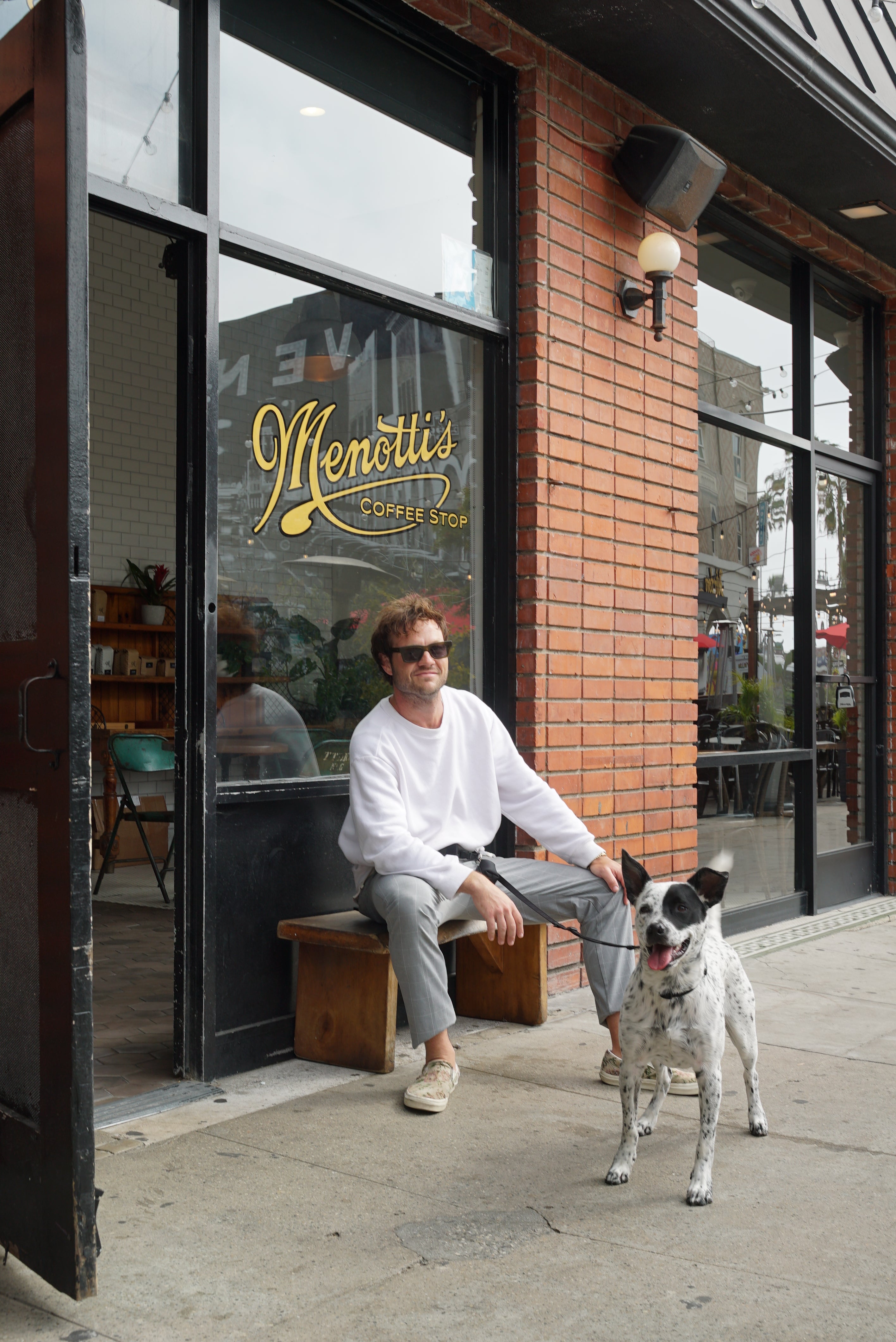 A  man wearing sunglasses and a white sweatshirt sitting on a wooden bench outside Menotti's Coffee Stop, holding the leash of a black and white dog standing next to him. The dog is on a leash from Lulu's from Cali. The man is looking towards the camera, and the coffee shop's logo is visible on the glass door.
