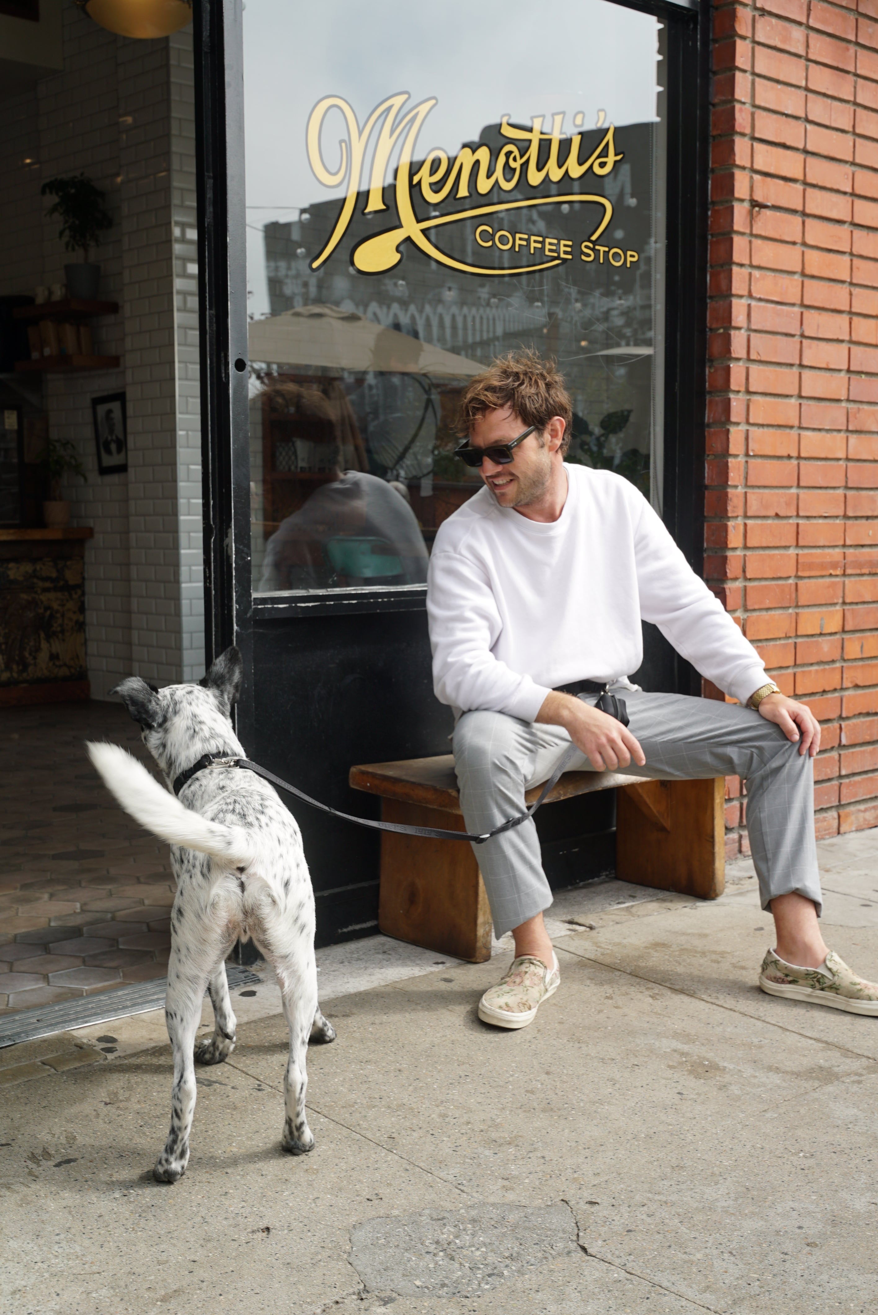 A man wearing sunglasses and a white sweatshirt sitting on a wooden bench outside Menotti's Coffee Stop, smiling at a black and white dog. The dog is on a leash from Lulu's from Cali and is looking towards the coffee shop entrance.