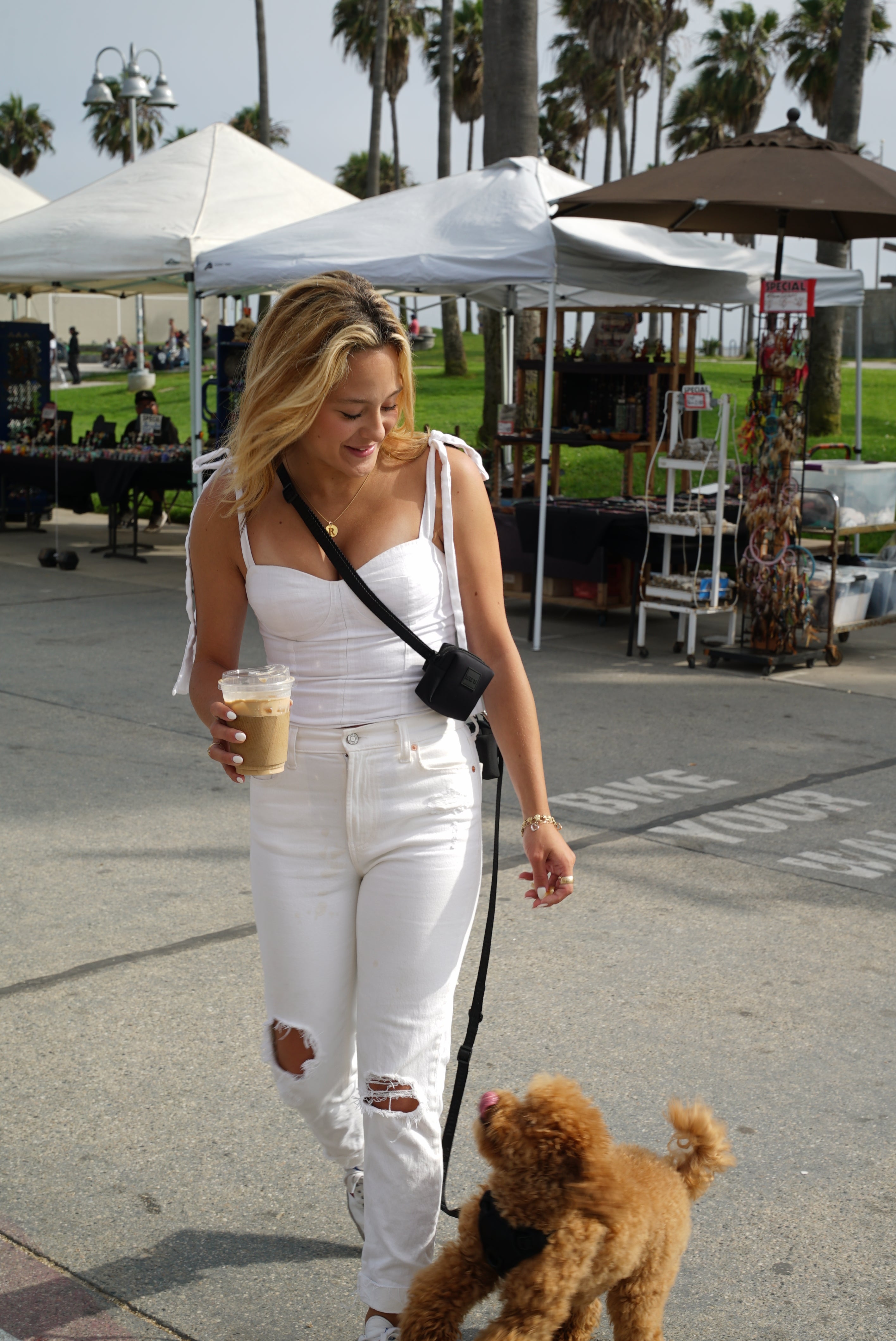 Blonde woman in a white outfit walking with a small brown dog on a hands-free leash in stealth black color from Lulu's from Cali, worn as a crossbody, at an outdoor market. She is holding a cup of coffee and smiling down at the dog