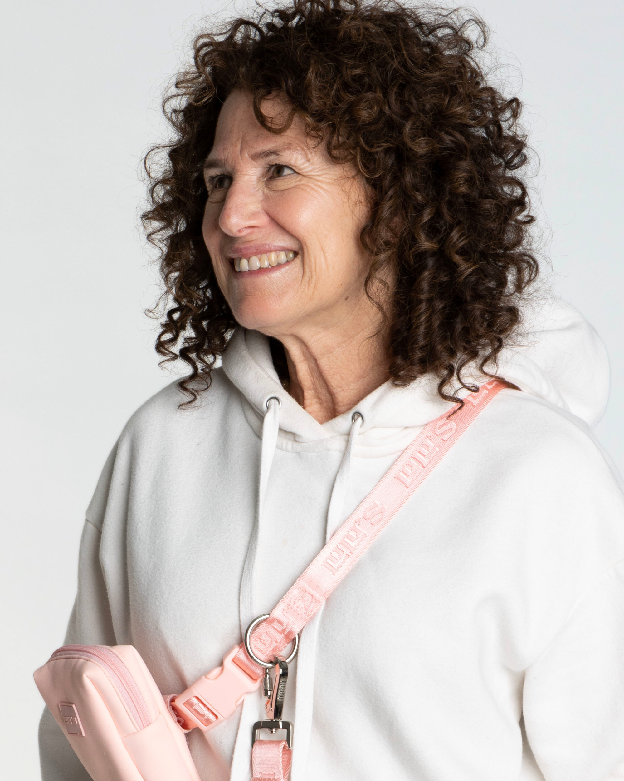 A woman wearing a white hoodie and a Blush pink Lulu's crossbody strap, attached to a matching Blush pink pouch. She is smiling and has curly hair.