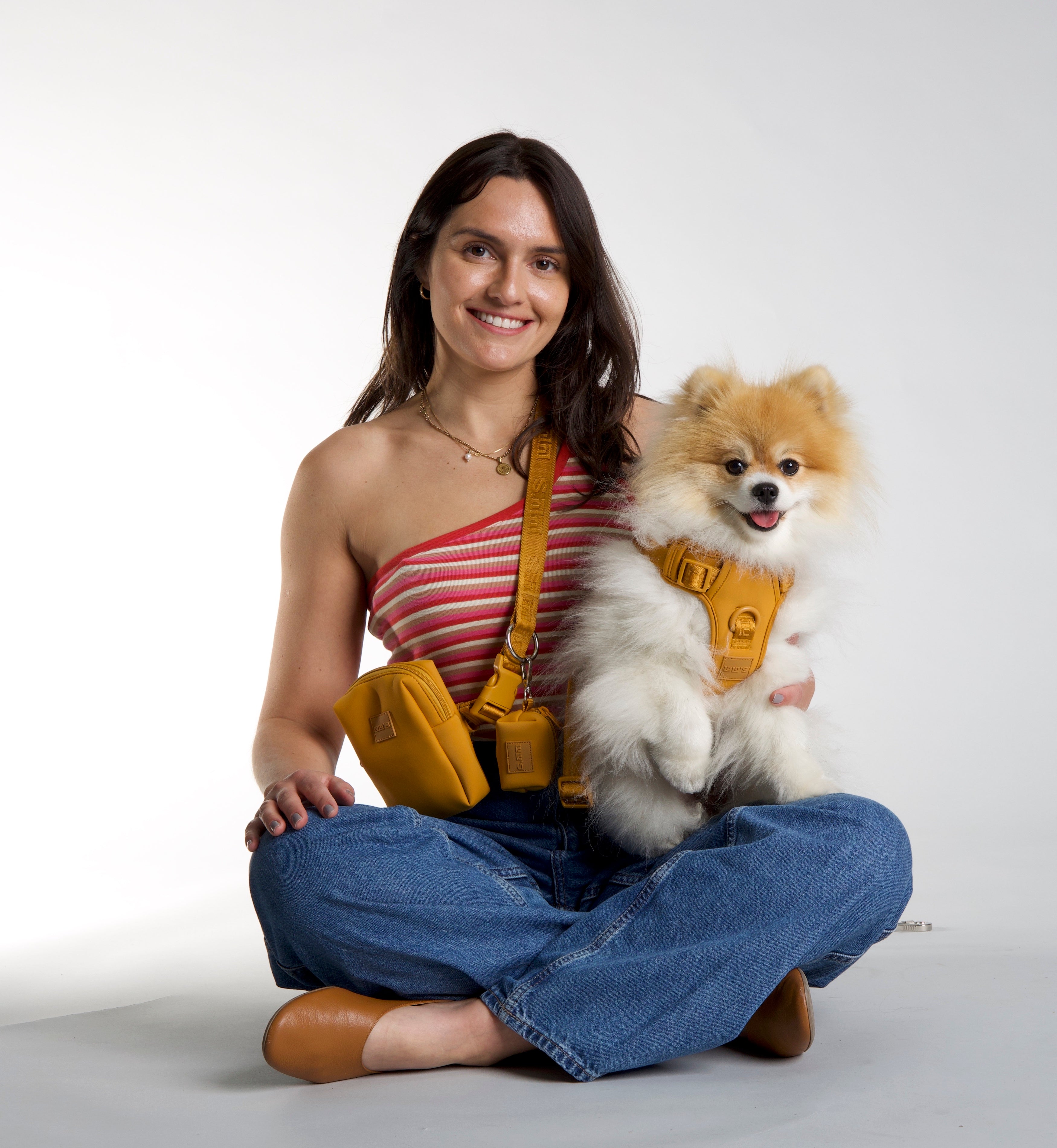 Smiling woman in a red striped top and jeans sitting cross-legged, holding a fluffy Pomeranian wearing a Sunburnt Orange harness from Lulu's